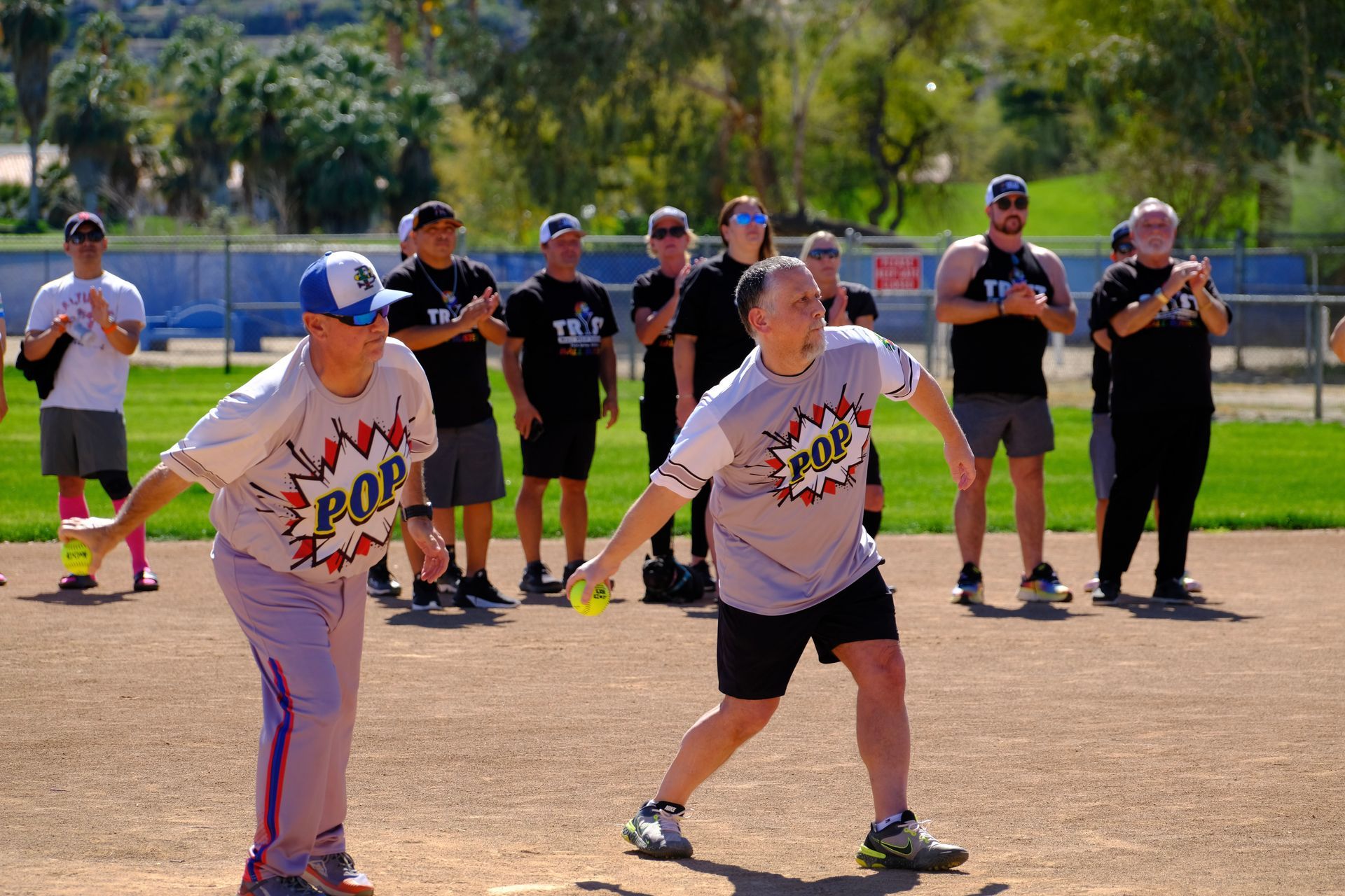 Two people in baseball jerseys stand ready on a dirt field, with a group of onlookers behind them.