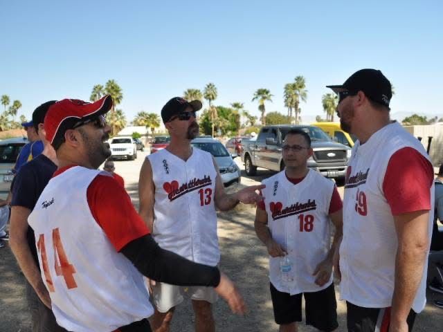 Four baseball fans in white-and-red jerseys chatting in a sunny parking lot before a game