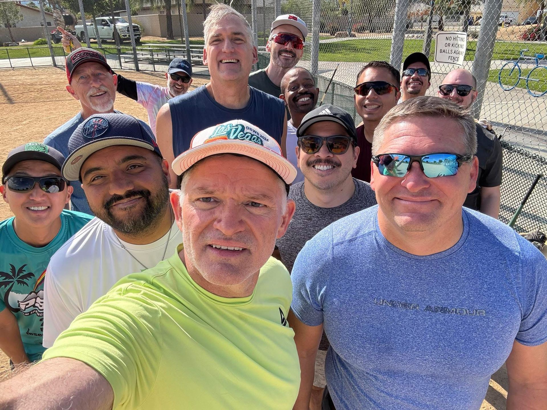 Group selfie of smiling friends outdoors in bright sunlight, with trees and a park behind them