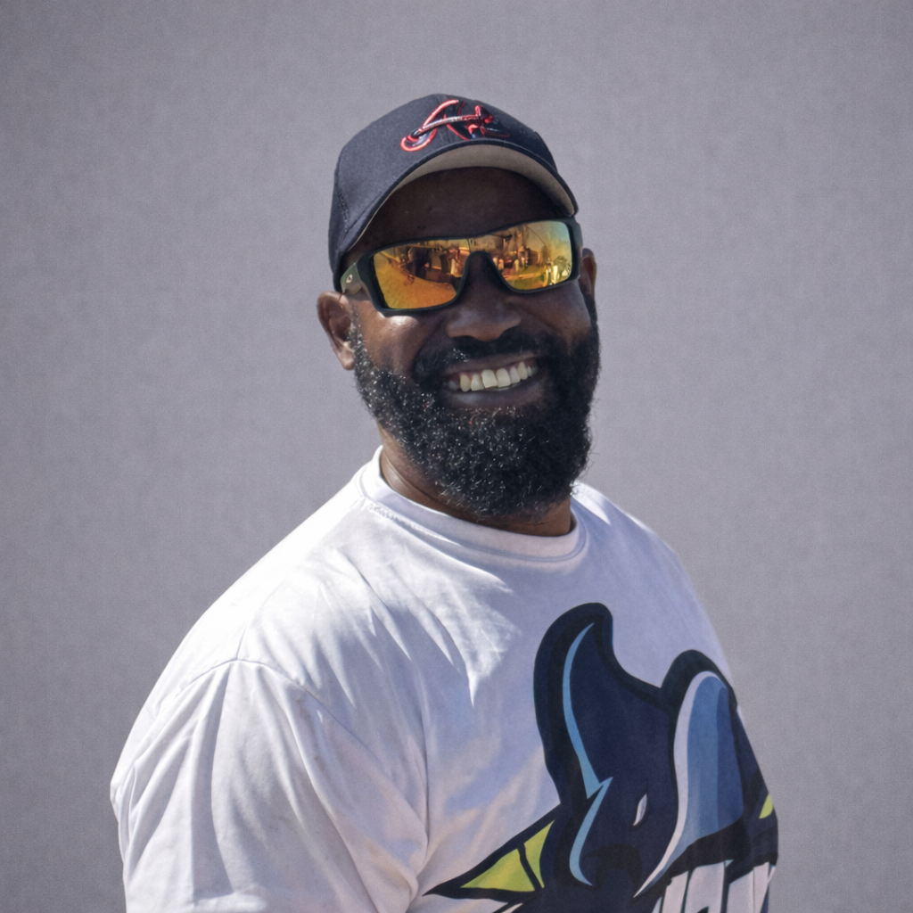 Smiling man in a black cap and sunglasses, wearing a white graphic T-shirt outdoors