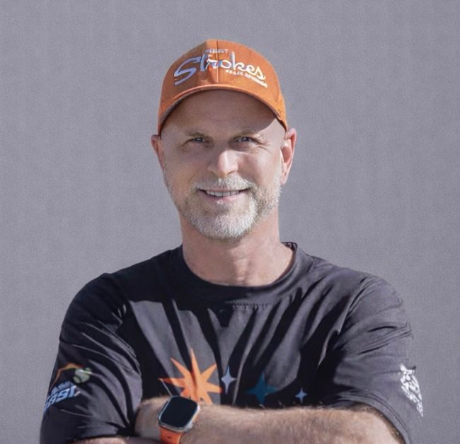Man wearing an orange cap and black T-shirt, smiling with arms crossed against a gray background