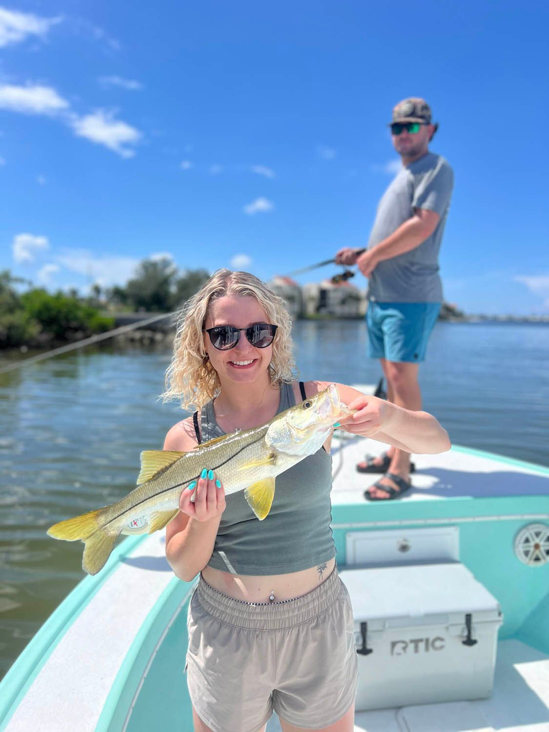A woman is holding a fish on a boat while a man is fishing in the background.