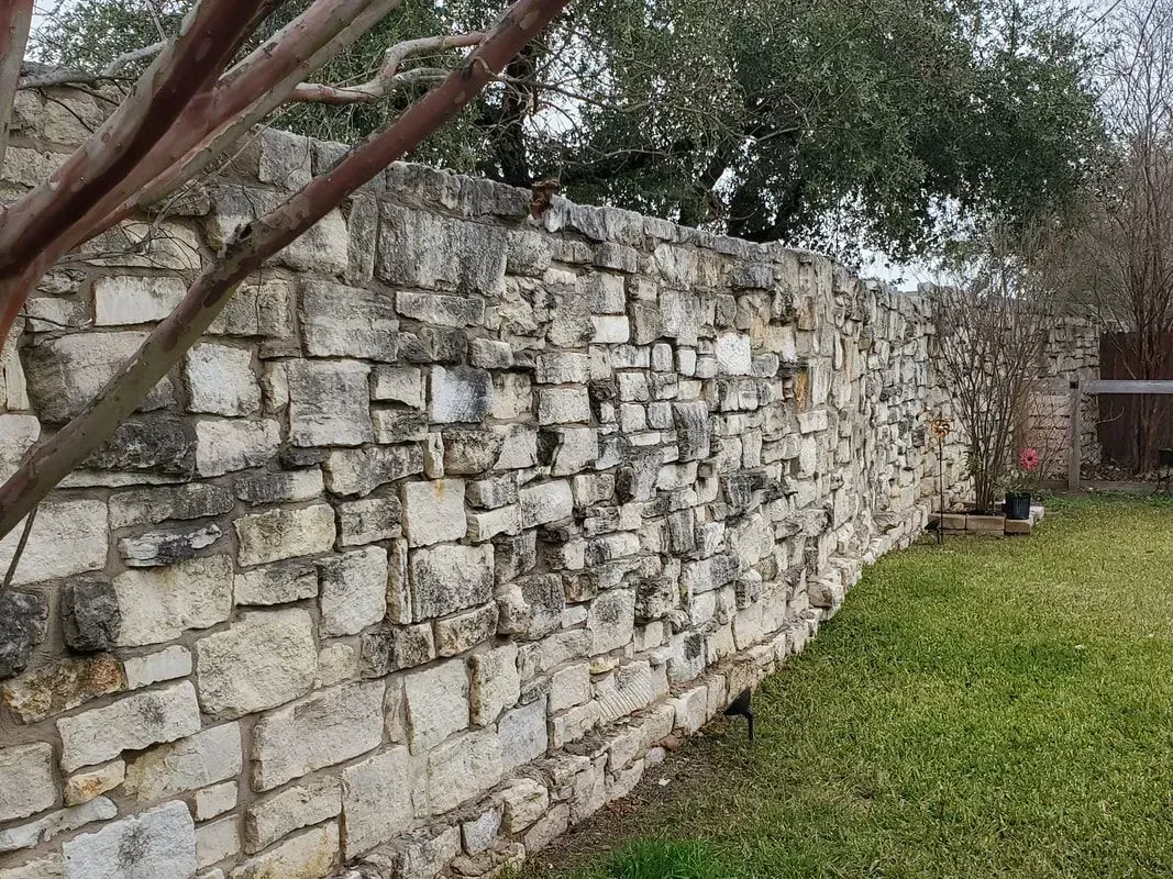 A large stone wall is surrounded by grass and trees in a backyard.