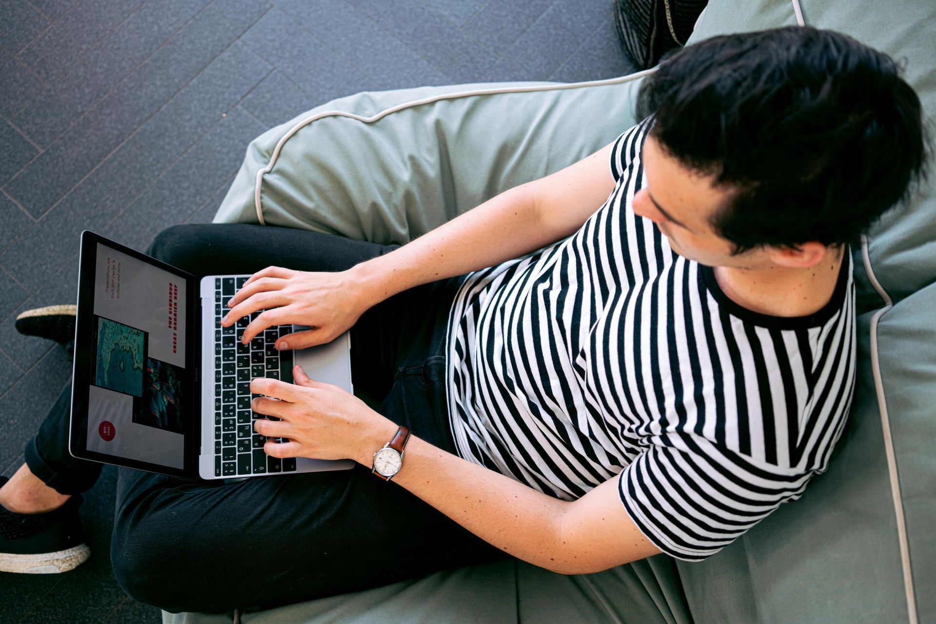 Person seated on a beanbag using a laptop; wearing striped shirt and watch; casual setting.