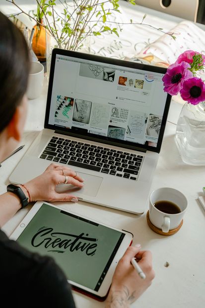 Person at a desk with a laptop and tablet, reviewing creative work, with coffee and flowers.