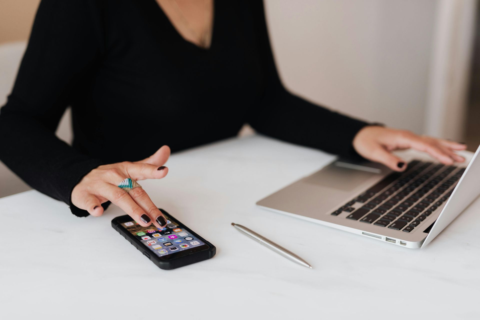 Woman using a smartphone and laptop at a desk, hands visible, black shirt.