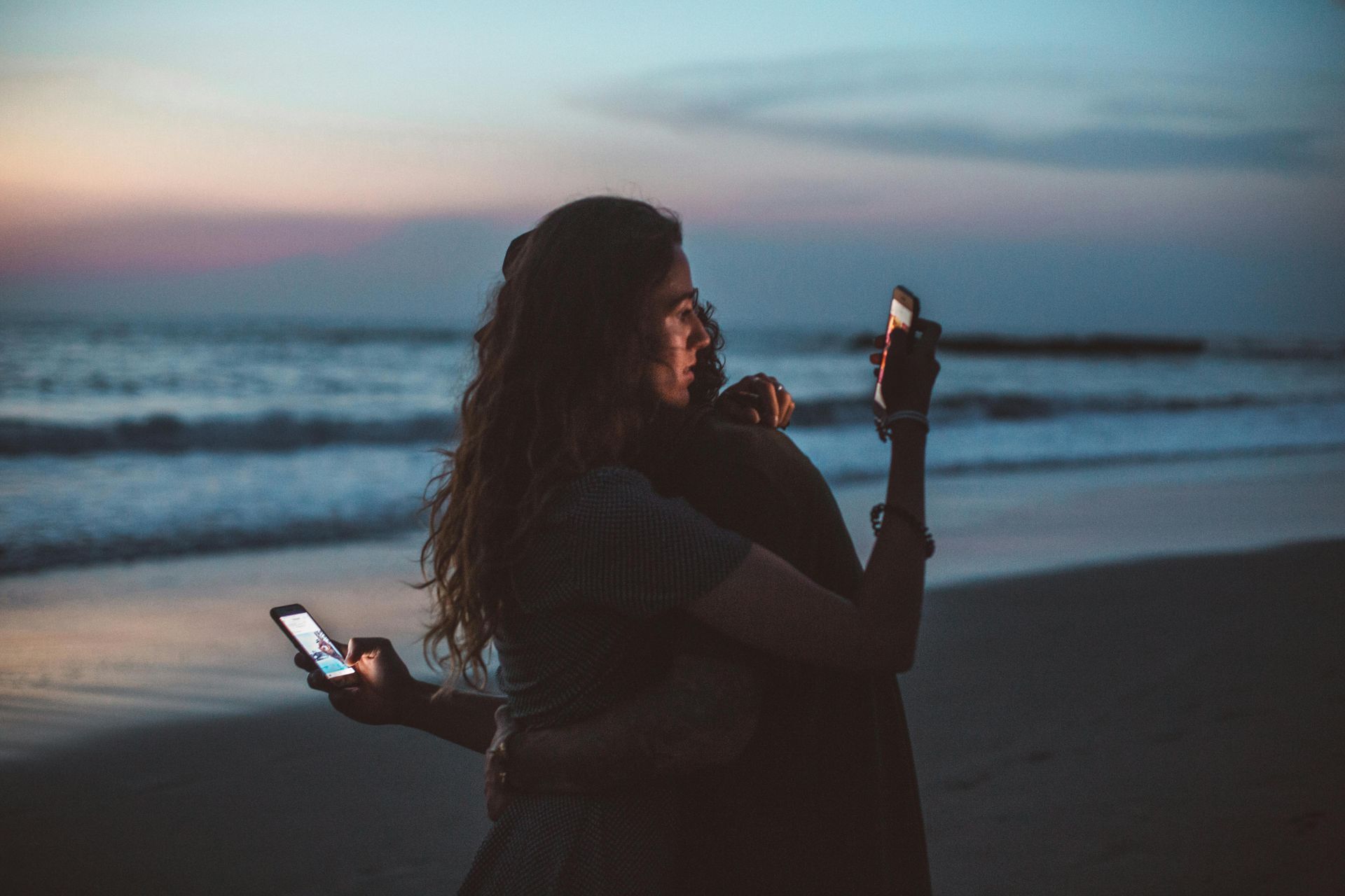 Two people hugging on beach, both holding illuminated phones, at sunset.