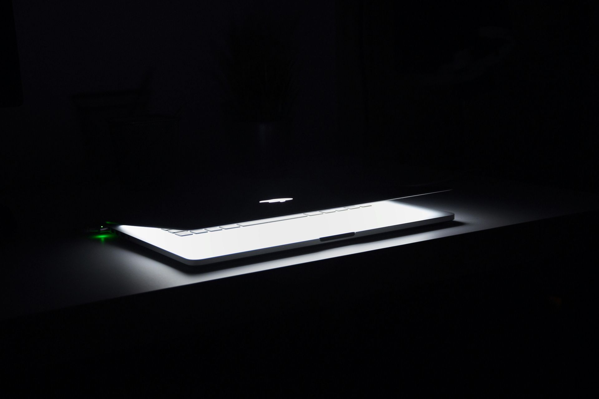 Laptop glowing brightly on a dark surface, corner with green light, in a dimly lit room.