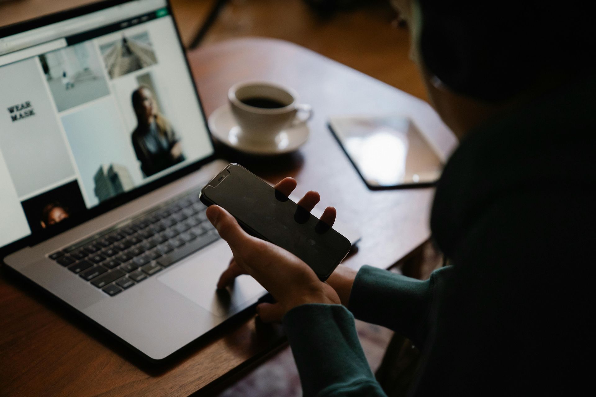 Person using laptop and phone at a table with coffee and tablet.