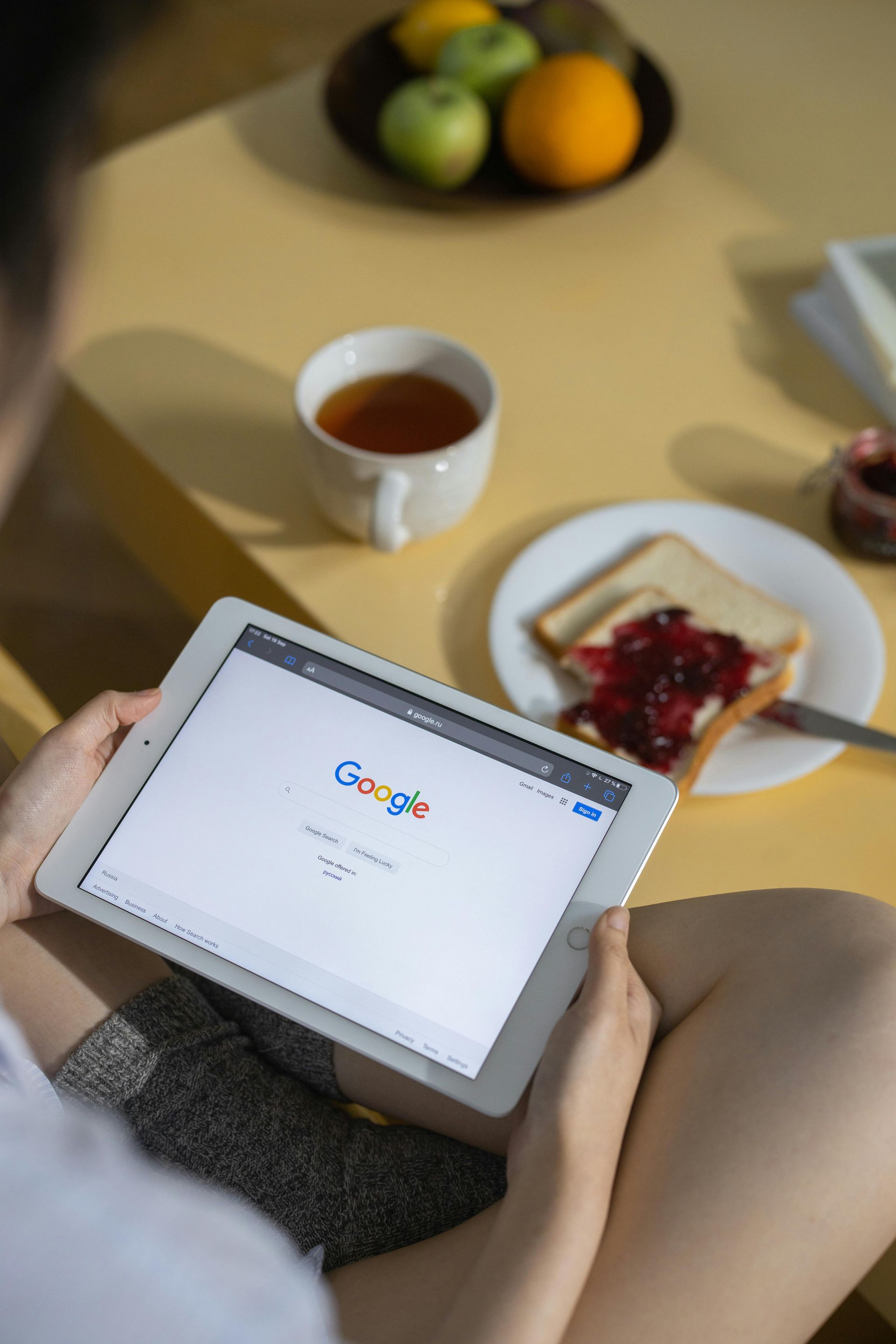 Person sitting with a tablet open to Google; breakfast with fruit, tea, and toast on the table.