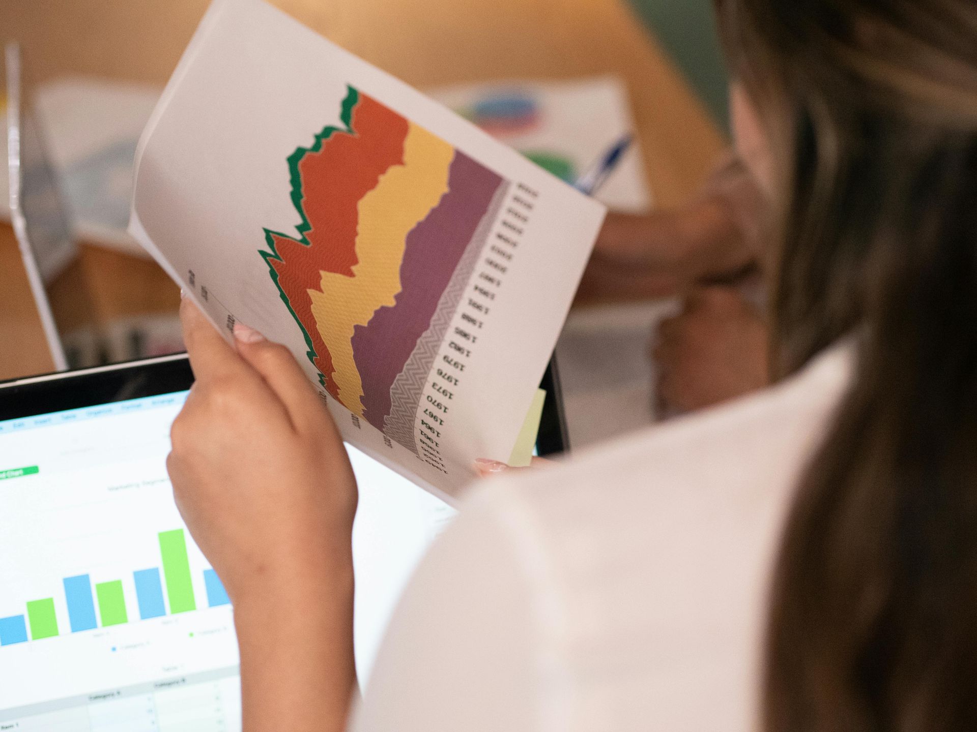 Woman analyzing charts, comparing paper data to laptop graph in bright office setting.