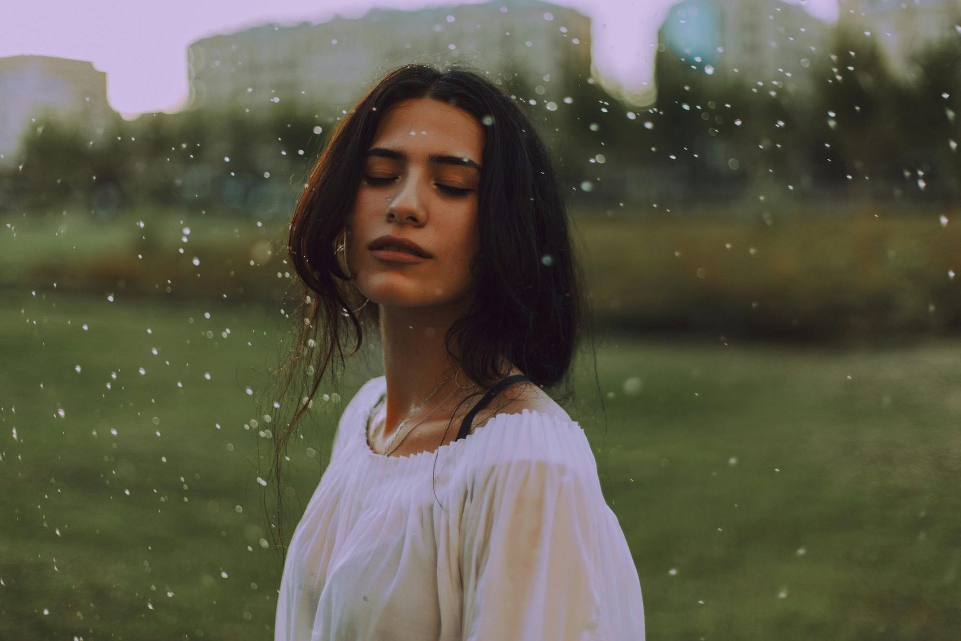 Woman with closed eyes in a white shirt, standing outside in the rain.