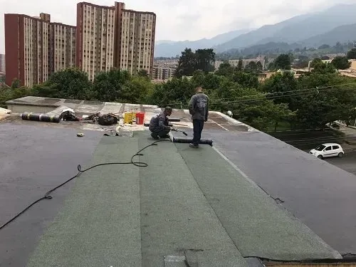 Trabajadores instalando techos en una azotea plana con edificios de la ciudad y montañas al fondo.
