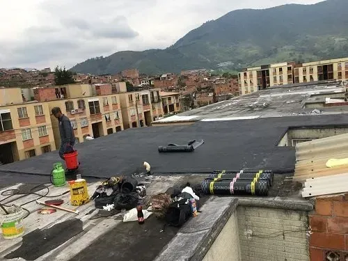 Trabajos de reparación en la azotea con fieltro asfáltico negro enrollado con vistas a edificios de apartamentos y montañas.