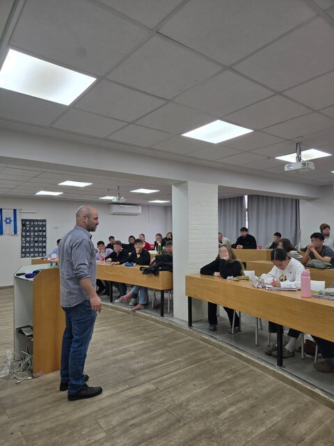 Man smiles at camera, another man writes near a laptop. Sunlight streams into the room.