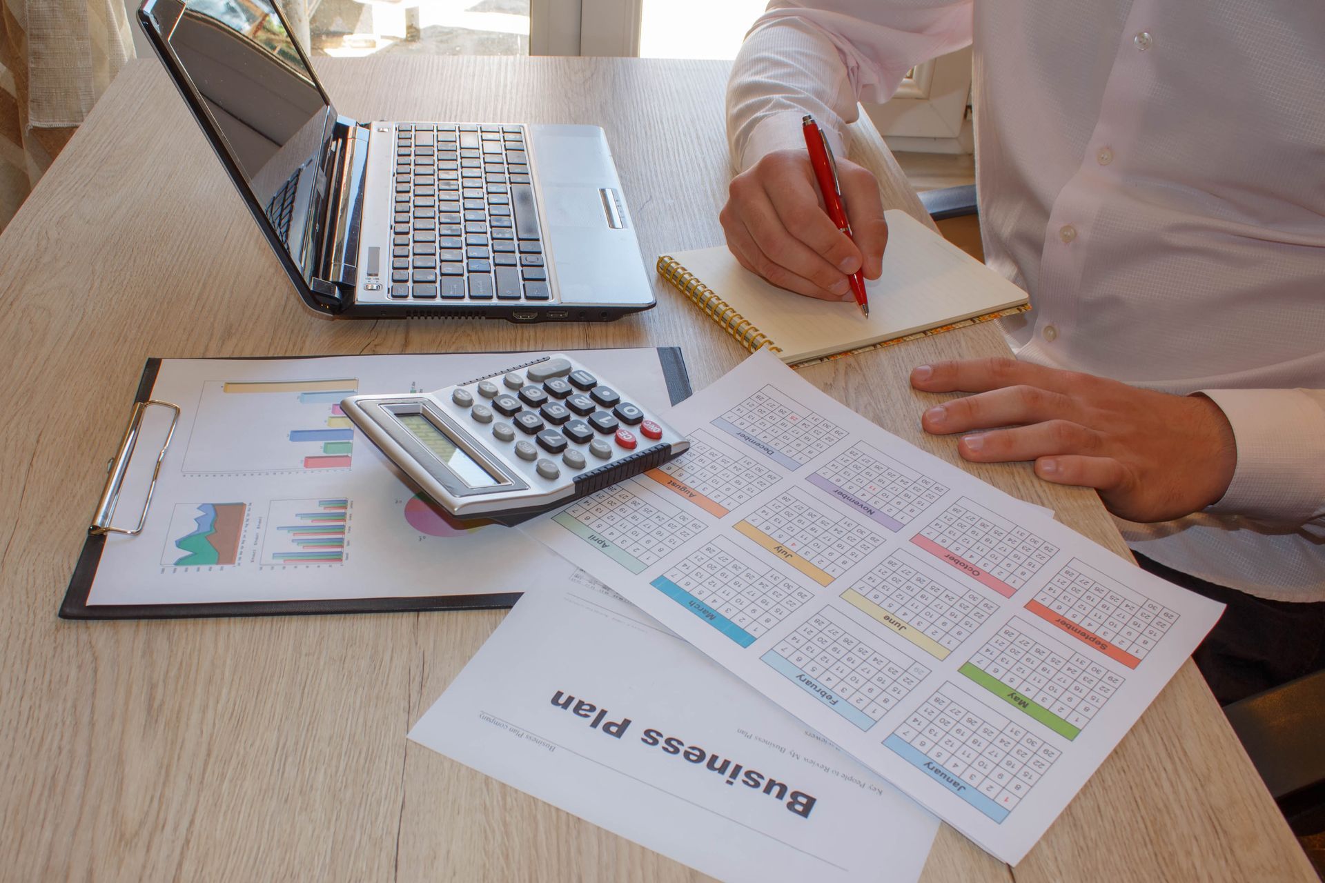 Person writing with red pen, surrounded by business documents, calculator, and laptop on a light wood desk.