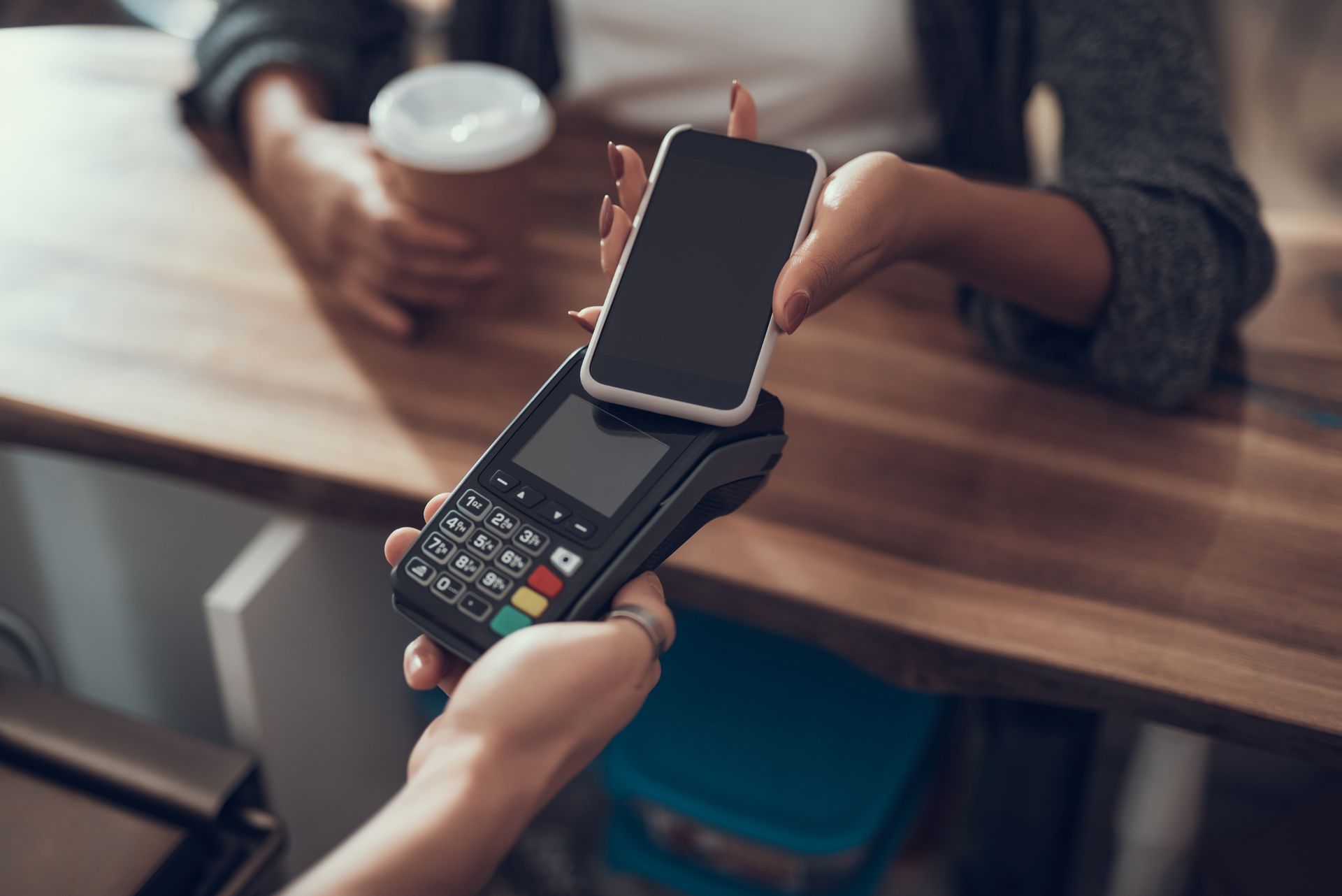 Person paying with phone at a point-of-sale terminal. A coffee cup is visible.