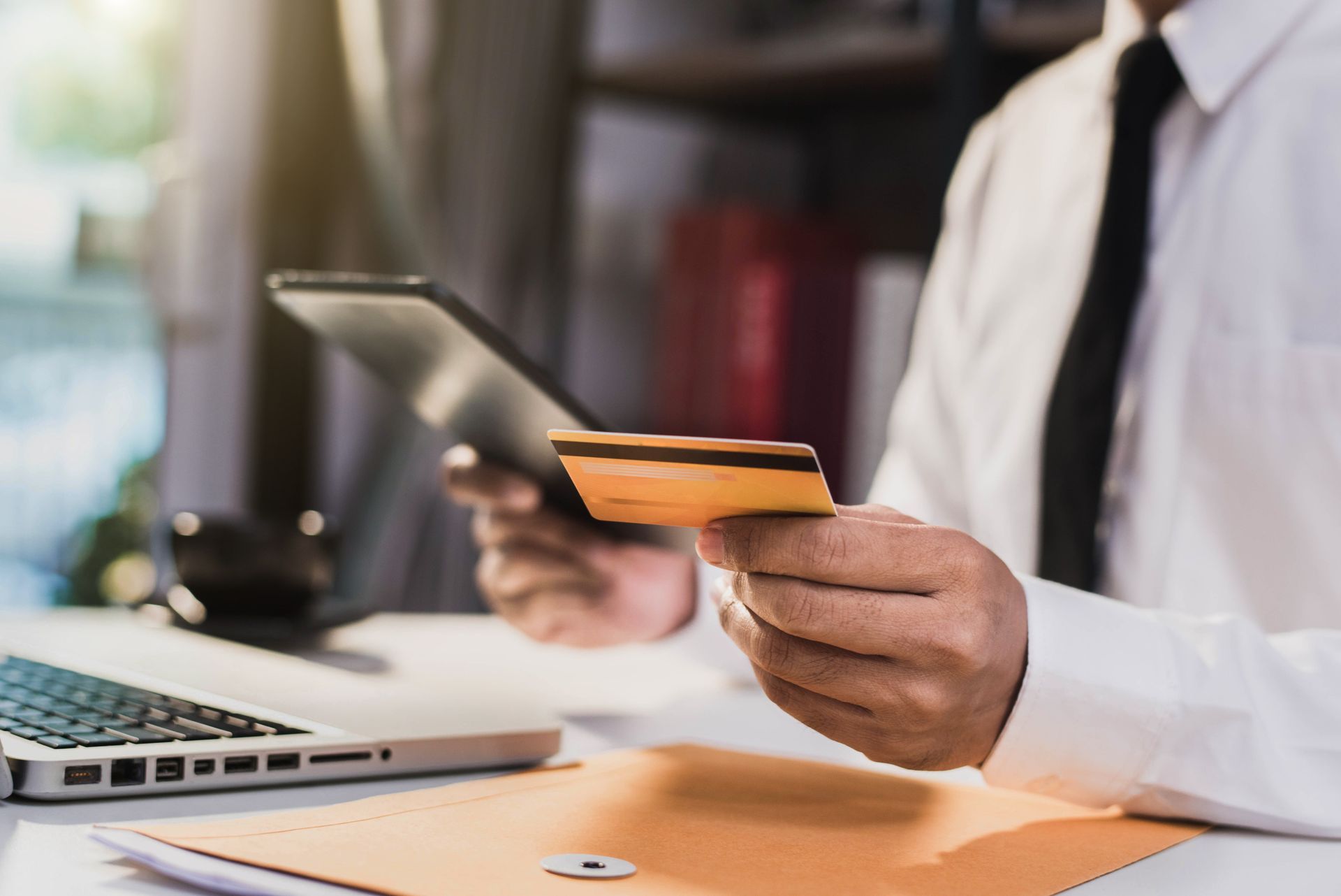 Person in office attire holding a credit card and tablet next to a laptop.