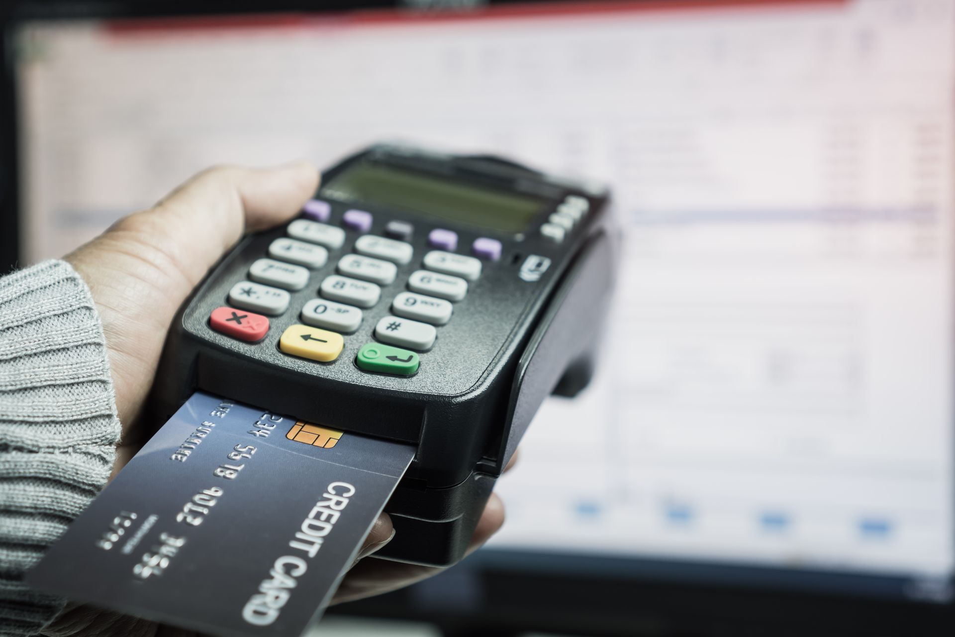 Person holding a credit card inserted into a black card reader with a computer screen in the background.
