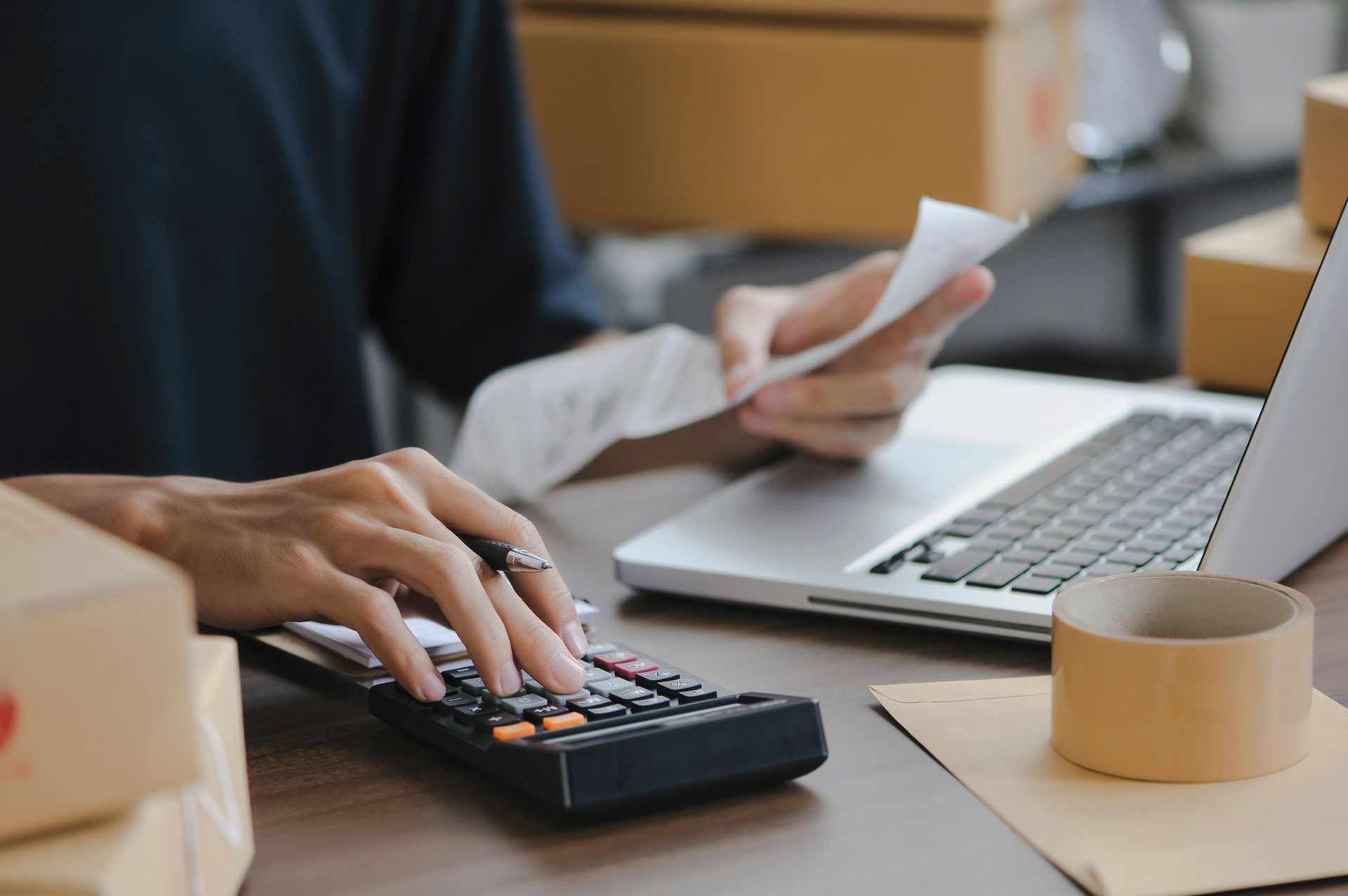 Person using a calculator, holding a receipt, and working on a laptop surrounded by shipping boxes.