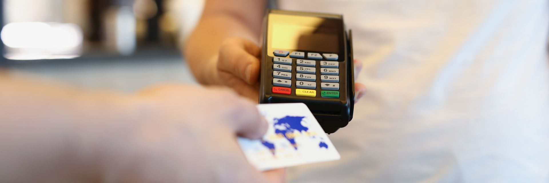 Hand holding a credit card over a point-of-sale device. The card has a world map design.