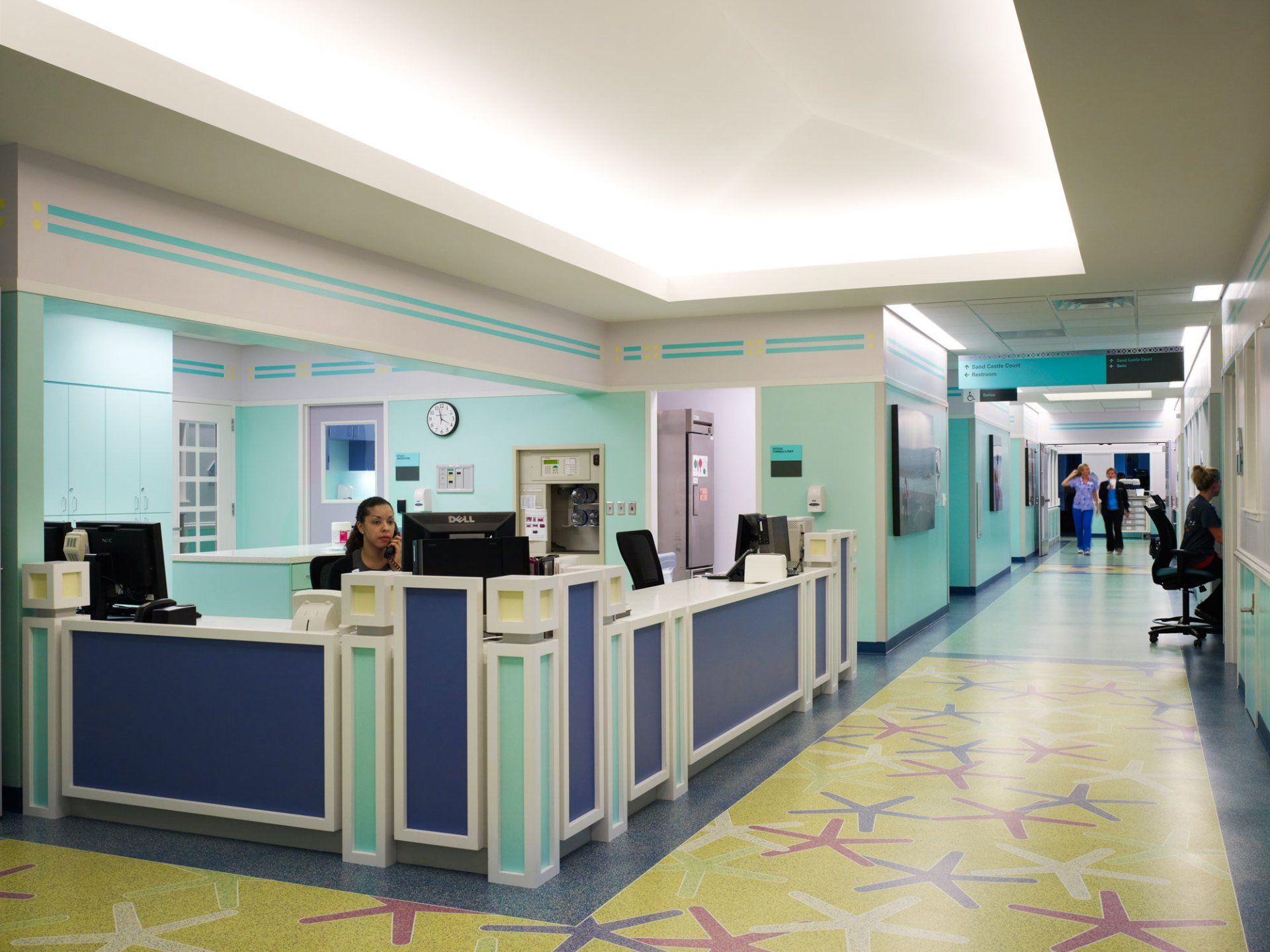 Hospital reception desk with a person behind it, looking at a computer. Hallway with patients and staff is visible.