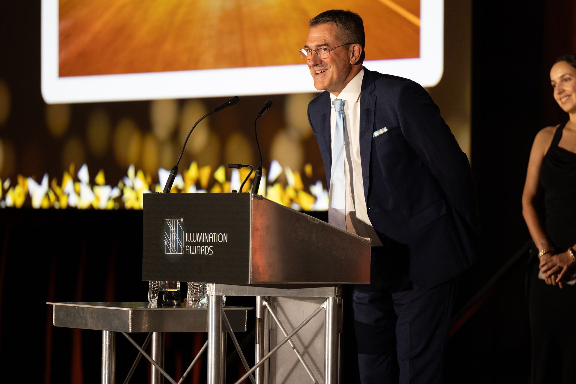 Man in a suit speaks at a podium. Woman stands to the side, smiling. Event backdrop.