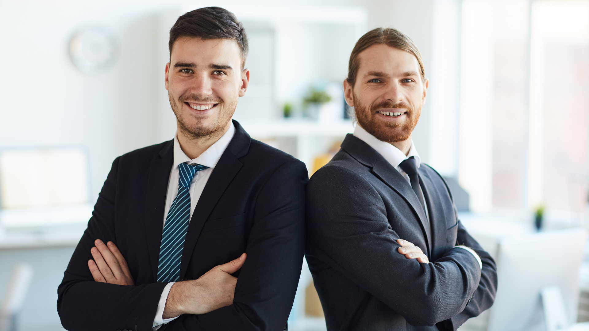 Two men in suits and ties are standing back to back with their arms crossed.