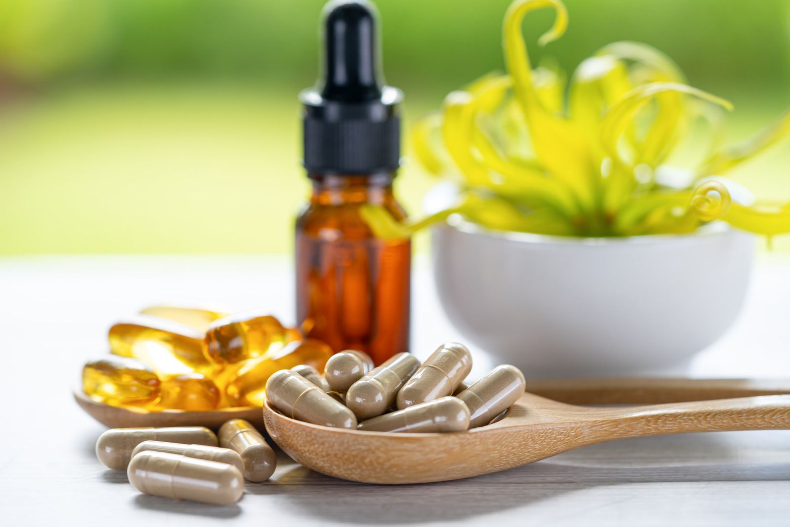 A wooden spoon filled with pills next to a bowl of pills and a bottle of oil.