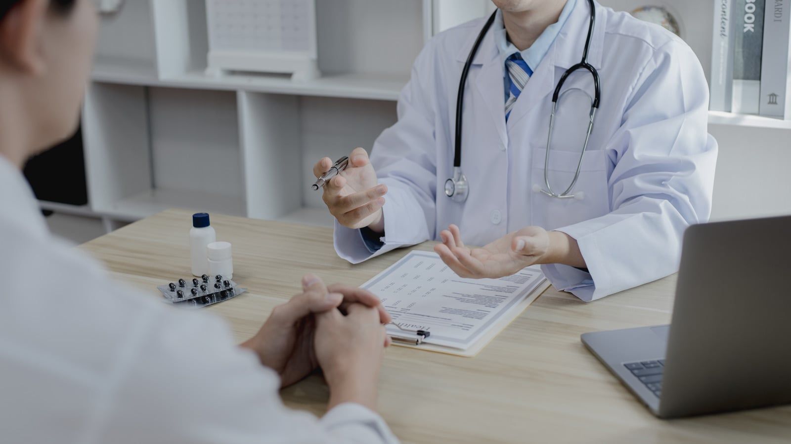Doctor consulting with a patient across a desk in a clinic, with a laptop and medical papers nearby.