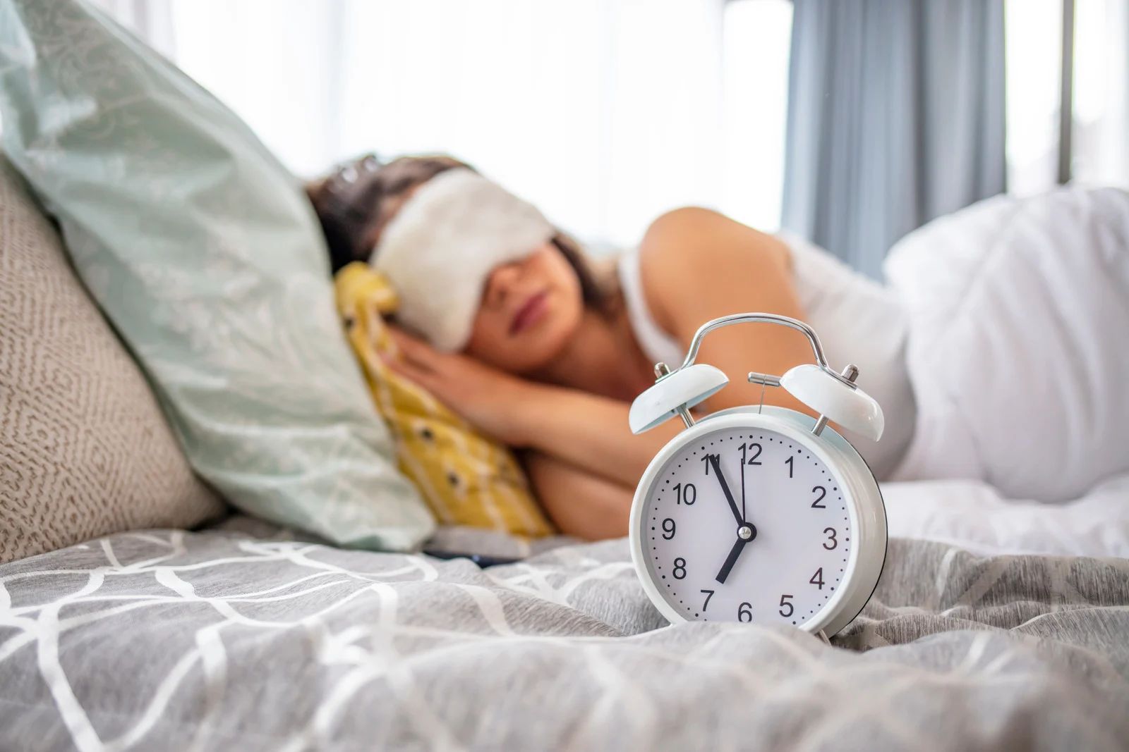 A person wearing an eye mask sleeps in bed with a white alarm clock set to seven o'clock in the foreground.