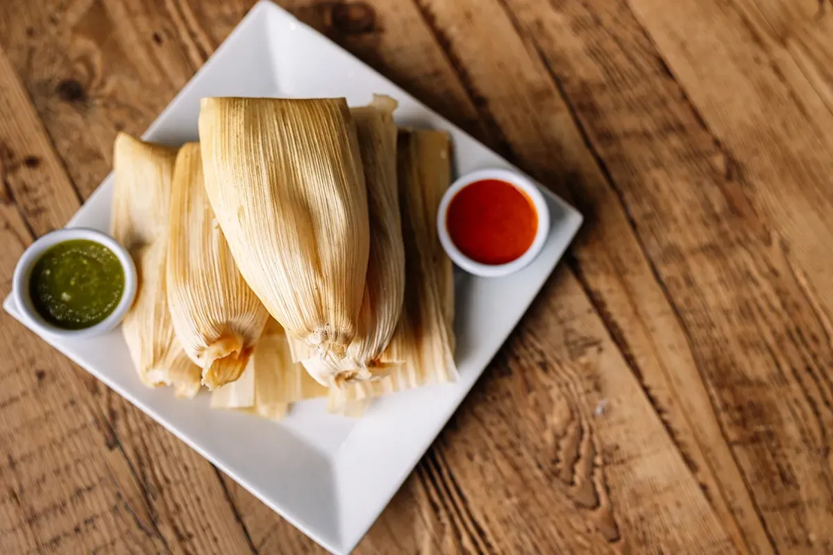 A white plate topped with tamales and two dipping sauces on a wooden table.