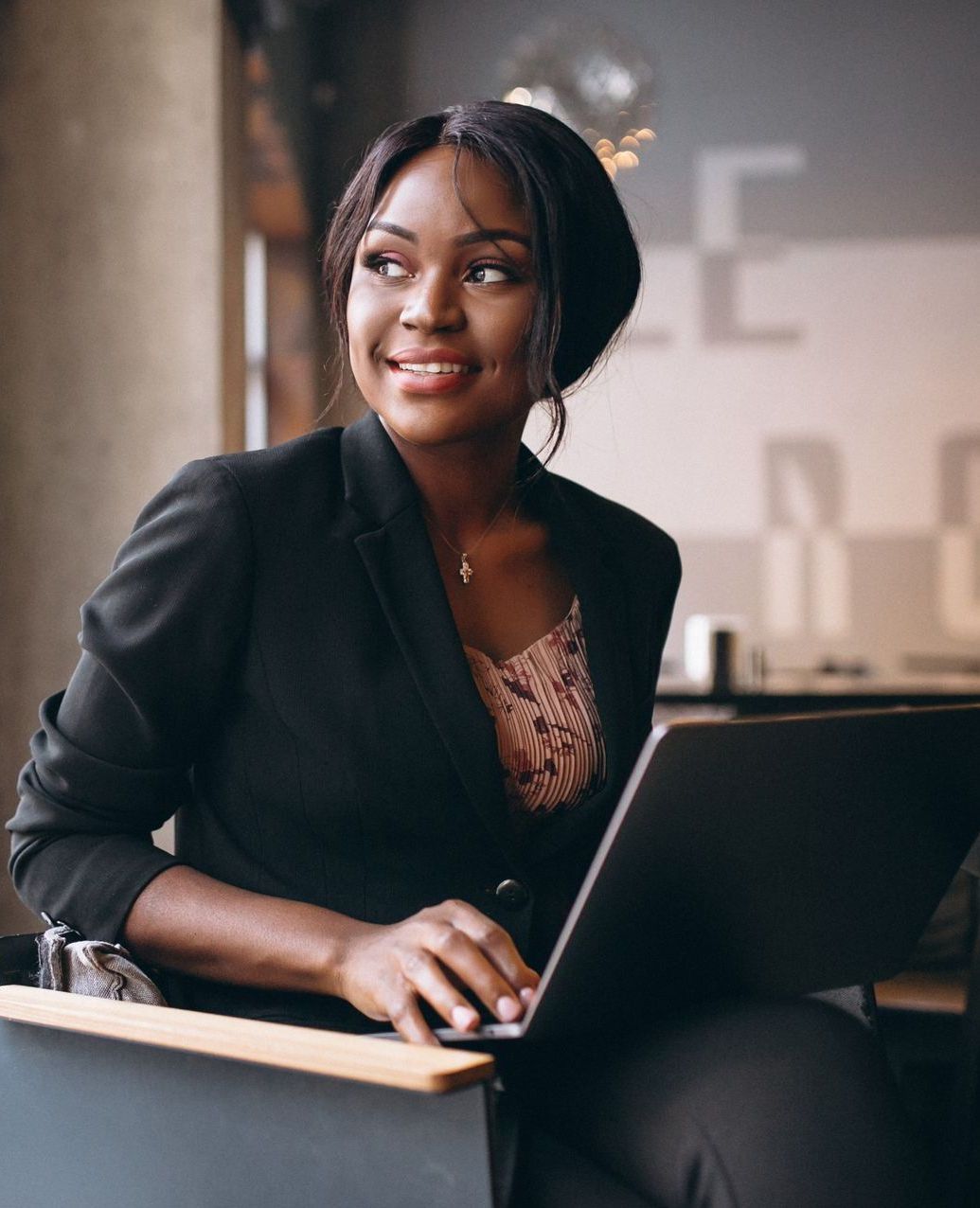 Woman in a black blazer smiles while using a laptop. Indoors, natural light.