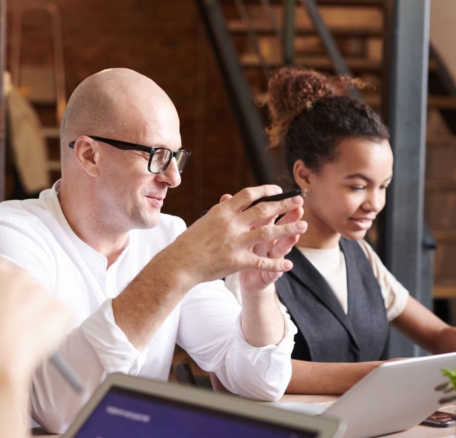Man with glasses gesturing, and woman working on laptop at a table, both smiling. Wooden stairs in background.
