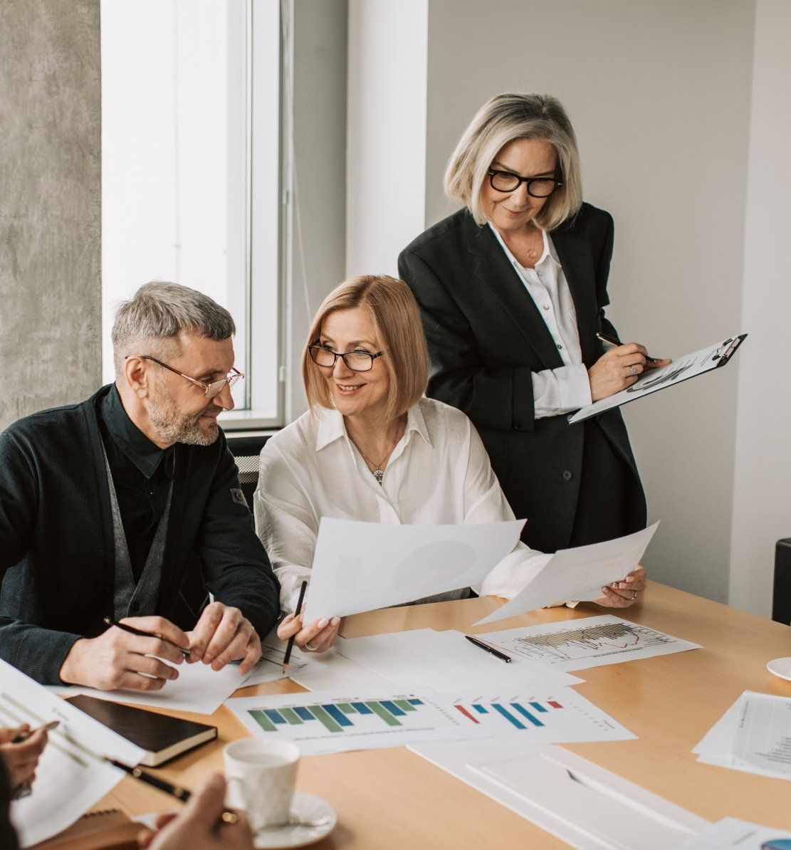 Business colleagues reviewing documents at a table. Charts and papers visible. One woman pointing.