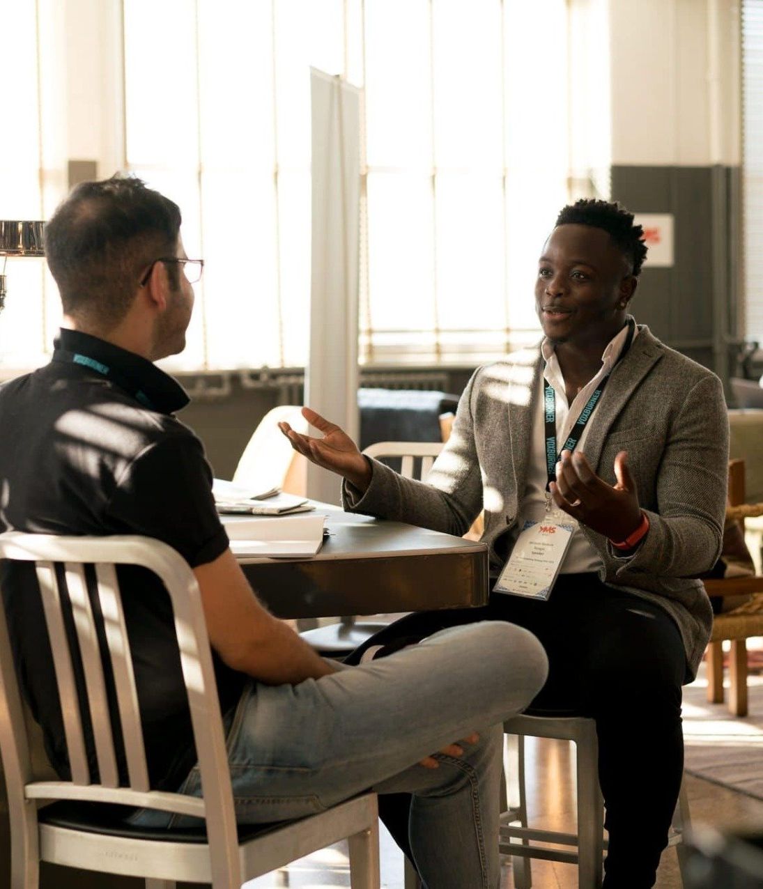 Two men seated at a table, conversing; one gestures with hands. Bright, sunlit setting.