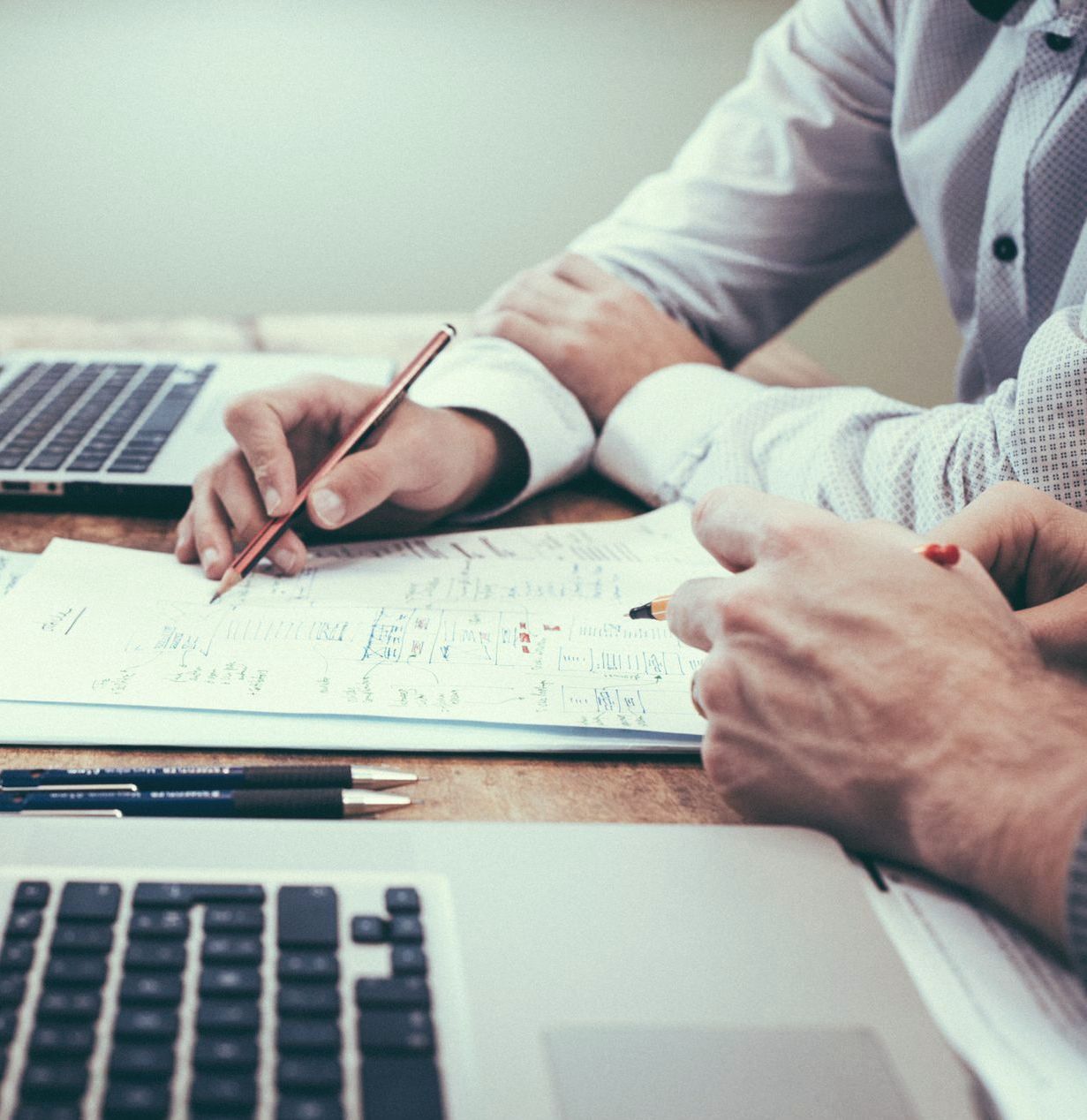 Two people in business shirts reviewing paperwork at a desk with laptops. One writes with a pencil.