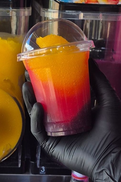 Hand holding a layered slushie in a clear cup: purple, red, orange, yellow.