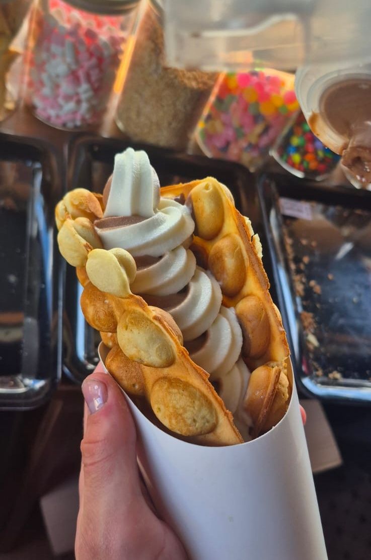 Hand holding a bubble waffle cone filled with soft serve ice cream and chocolate sauce.
