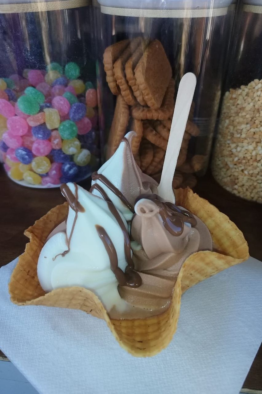 Soft-serve ice cream swirl in waffle cone bowl, drizzled with chocolate, candy and cookies in background.