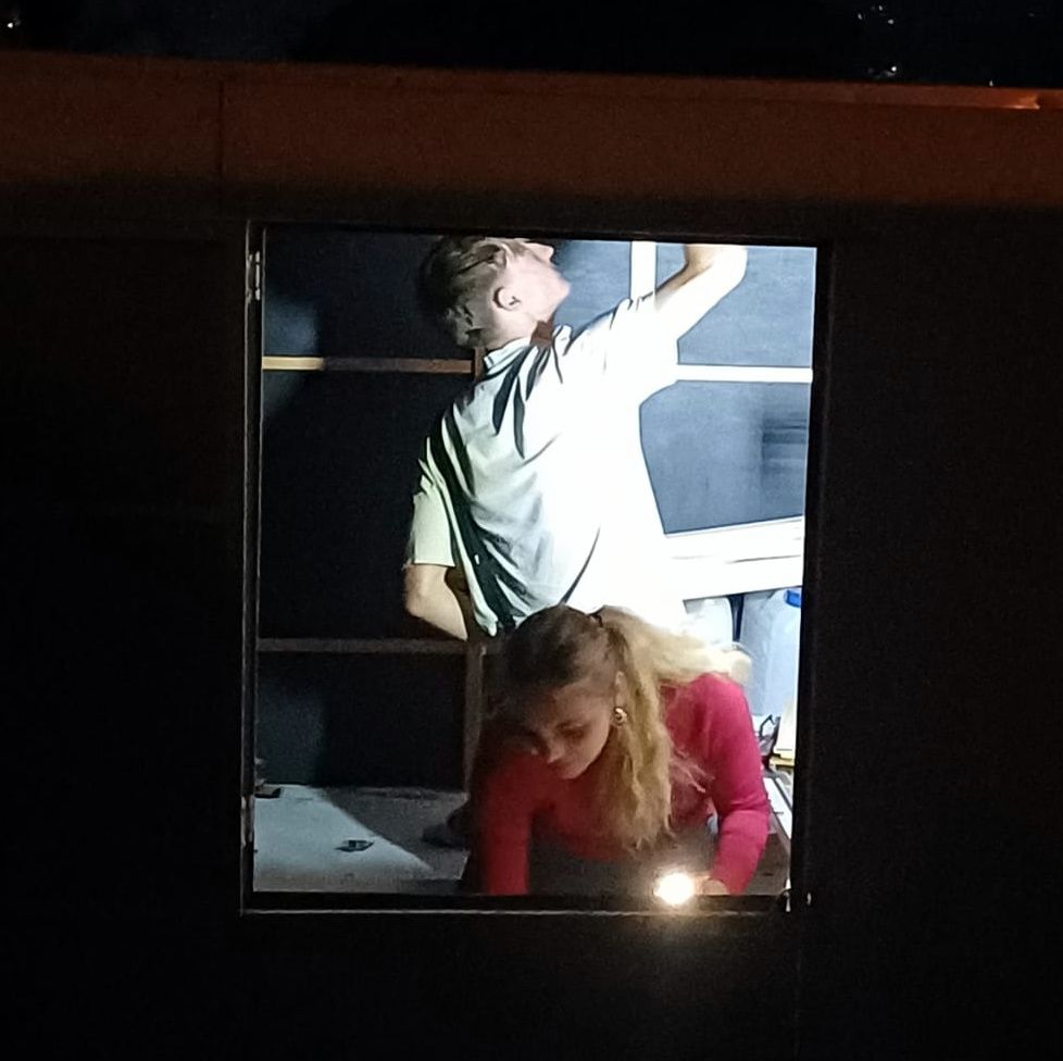 Two people in a dark room seen through a window. One stands, reaching up, the other leans down.