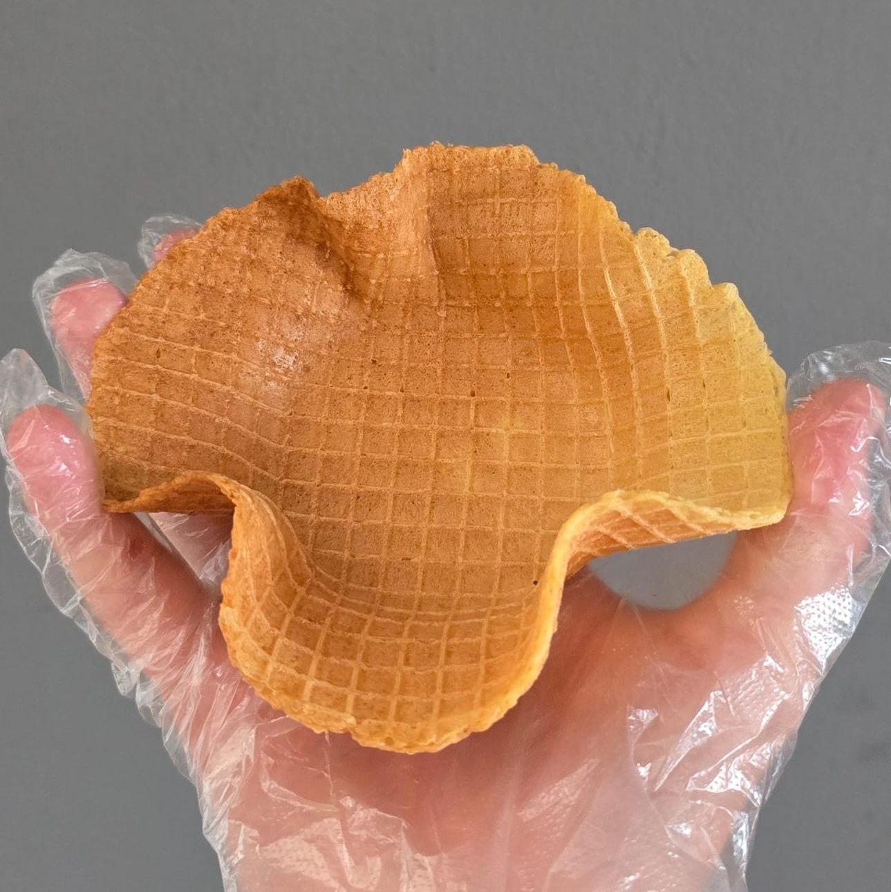 A hand wearing a clear glove holding a golden-brown waffle bowl.