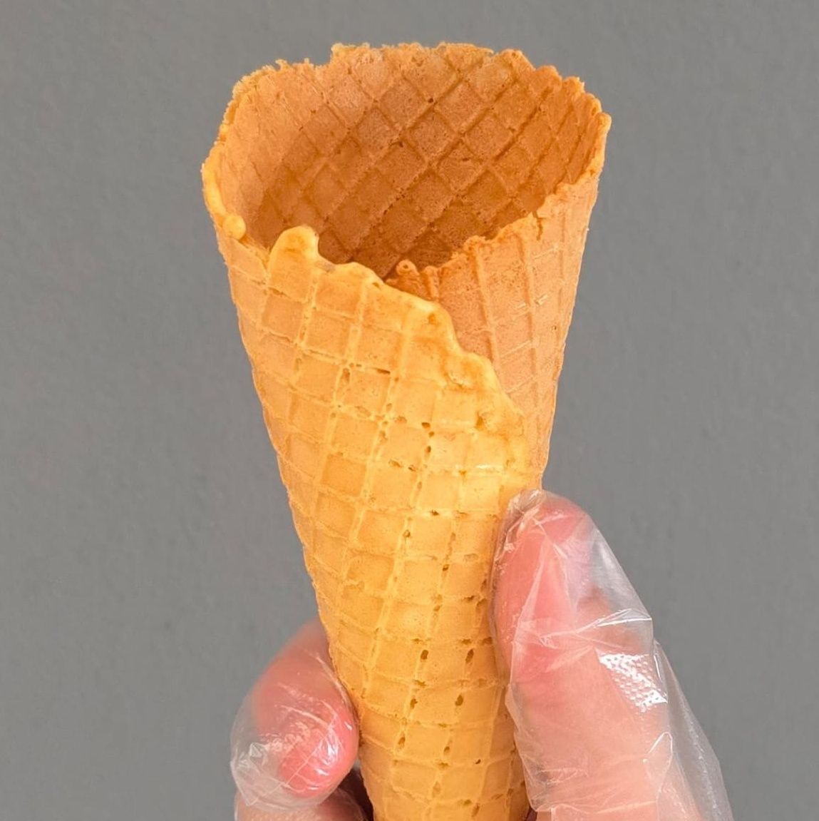 Hand holding an empty waffle cone against a gray background.