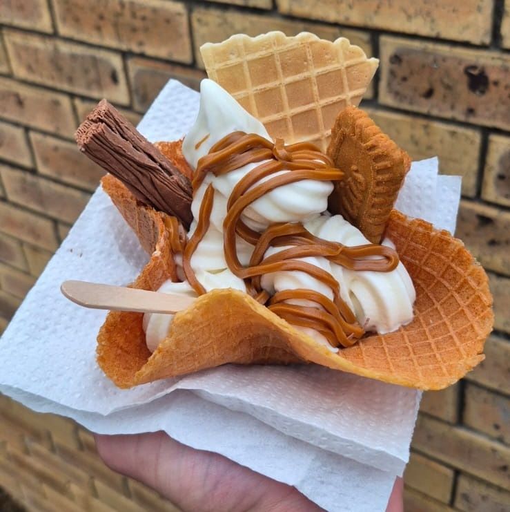 Soft serve ice cream in waffle bowl, drizzled with sauce, surrounded by wafers and chocolate bar. Held over a brick wall.