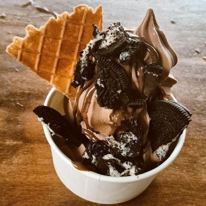 Chocolate soft serve ice cream with Oreo toppings and waffle cone in a white cup.