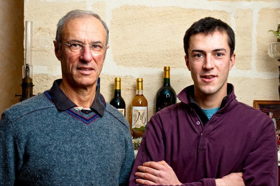 Deux hommes se tiennent l'un à côté de l'autre devant un mur avec des bouteilles de vin dessus.