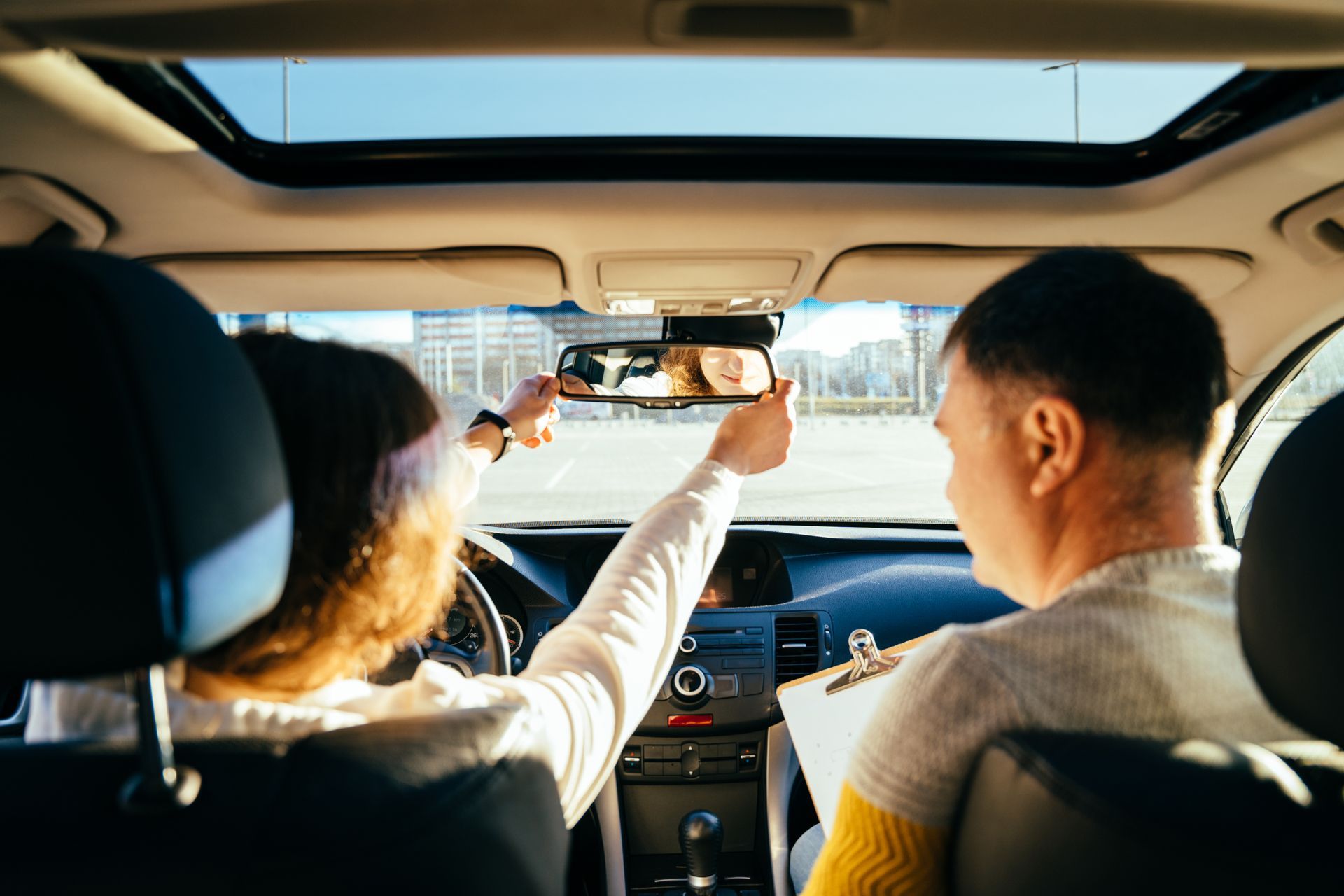 Person adjusting rearview mirror, sitting with instructor in car; clear sky.