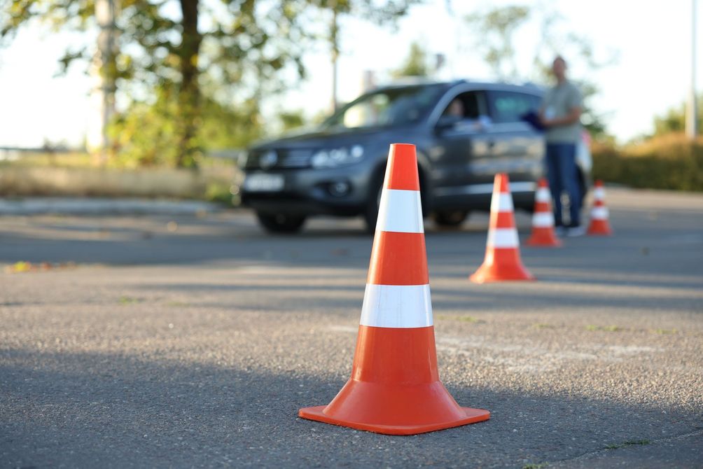 Orange and white traffic cones set up for driving test; car and instructor in background.