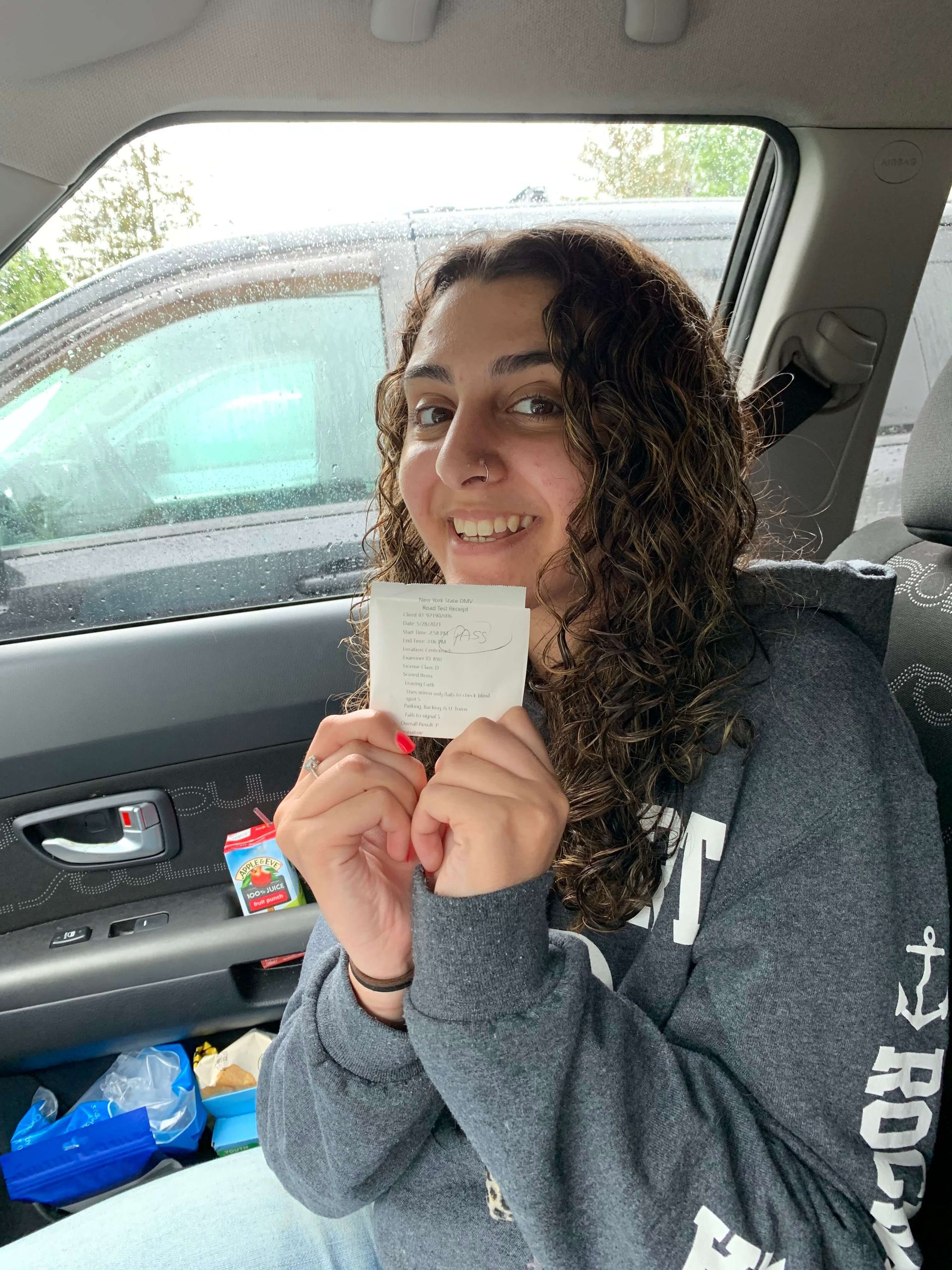 Woman in car smiles, holding receipt. Gray hoodie, curly hair.