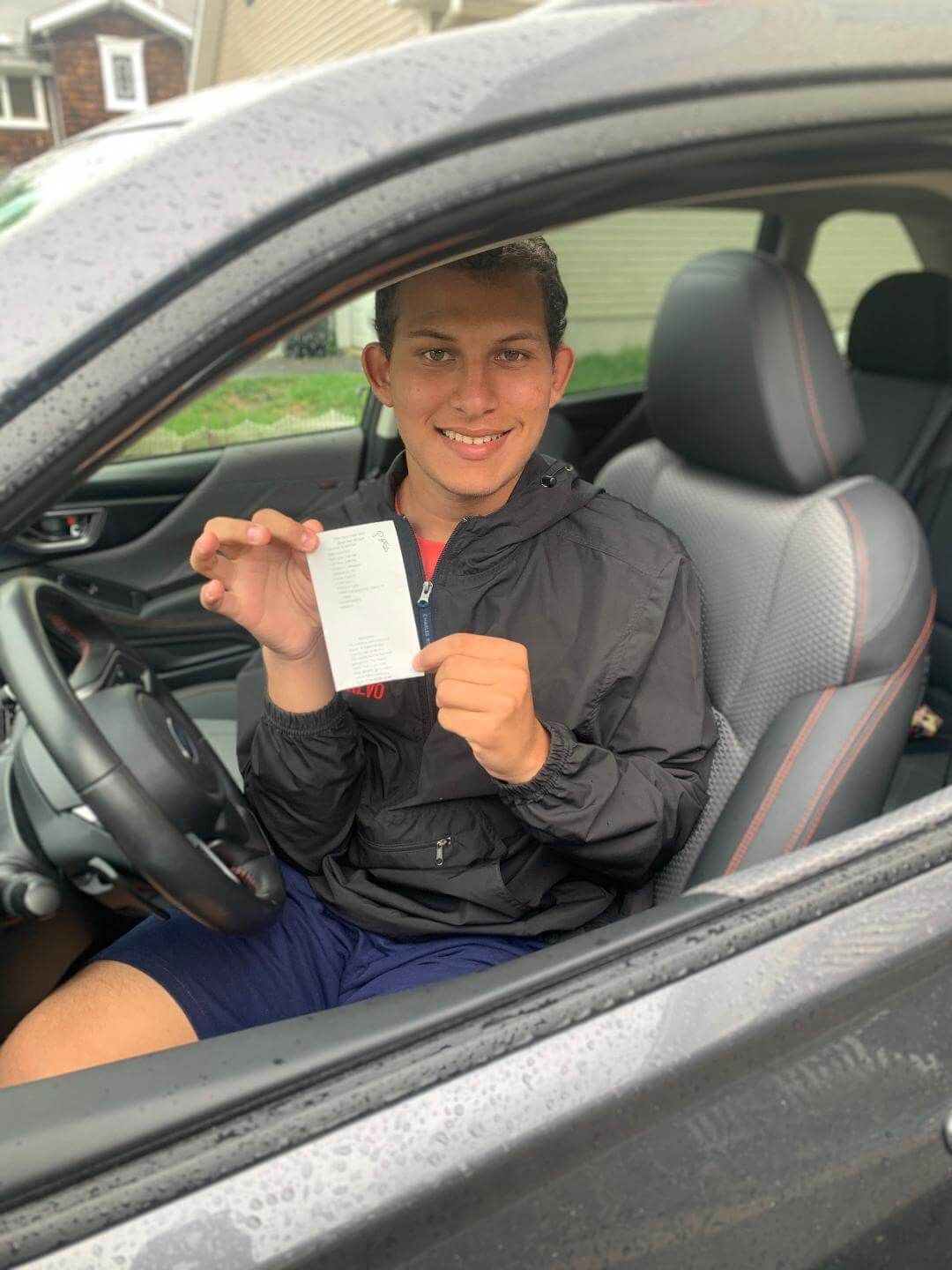 Person in a car holding a receipt, smiling. Dark car interior, cloudy outdoor setting.